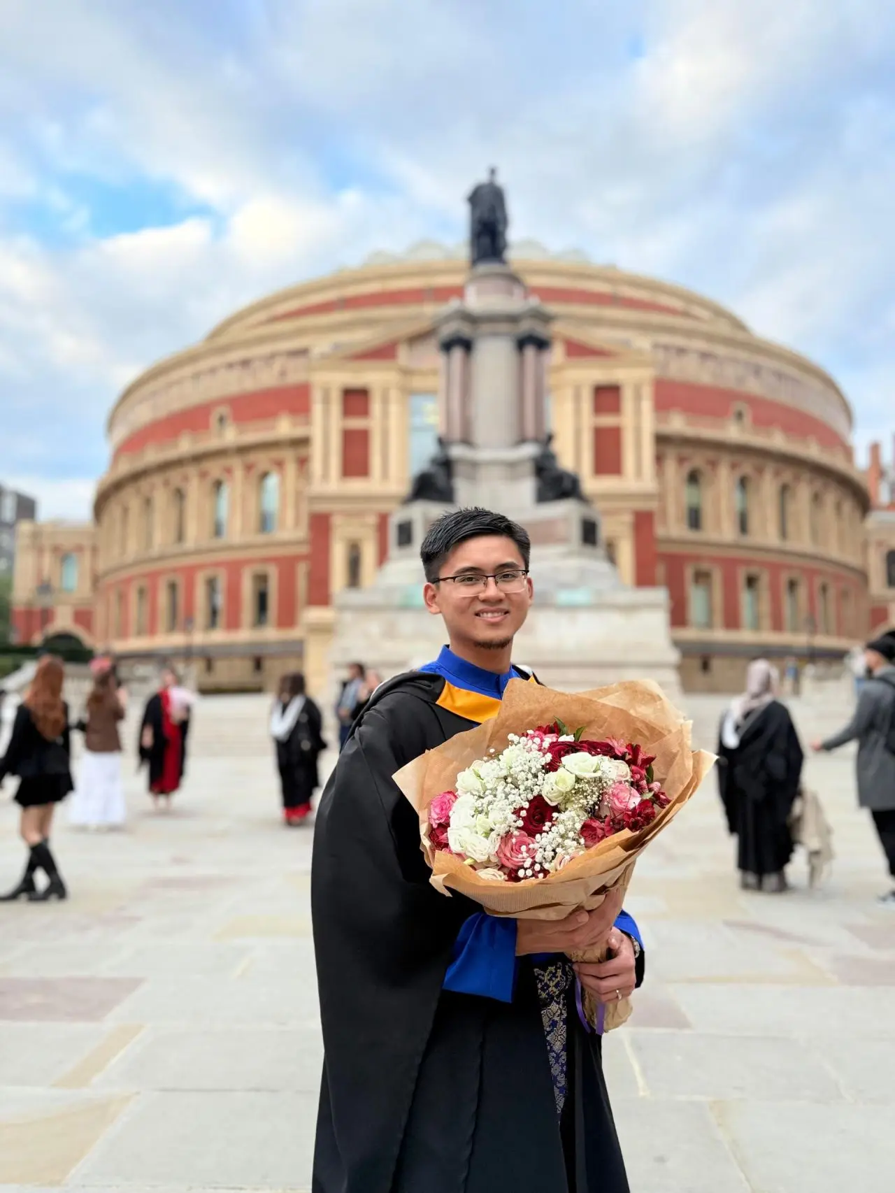 Hilman Azmi, anak kelahiran Terengganu, bergambar memakai jubah graduasi sambil memegang sejambak bunga di hadapan Royal Albert Hall semasa majlis konvokesyen Imperial College London.