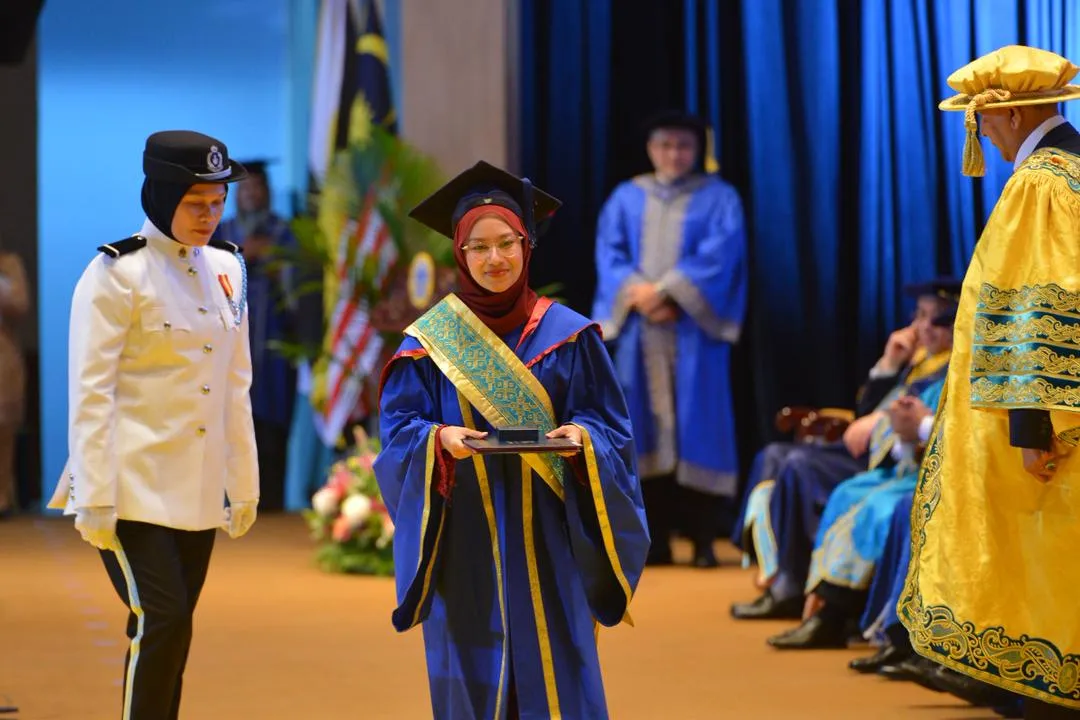 Graduan wanita berbaju jubah konvokesyen menerima anugerah di pentas majlis graduasi UUM, sambil diiringi pegawai protokol.