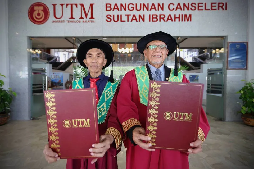 Dua graduan senior UTM bergambar dengan skrol di hadapan Bangunan Canseleri Sultan Ibrahim.