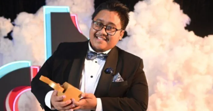 Abdul Qayyum Halid, pencipta kandungan (content creator) 'Che Sayang Kitchen', bergambar sambil tersenyum dan memegang anugerah di atas pentas. Beliau memakai tuksedo hitam dengan bowtie biru gelap, cermin mata, dan berlatarbelakangkan hiasan awan asap putih dan logo TikTok. Imej ini diambil semasa majlis TikTok Awards 2024 di mana beliau dinobatkan sebagai Creator of The Year.
