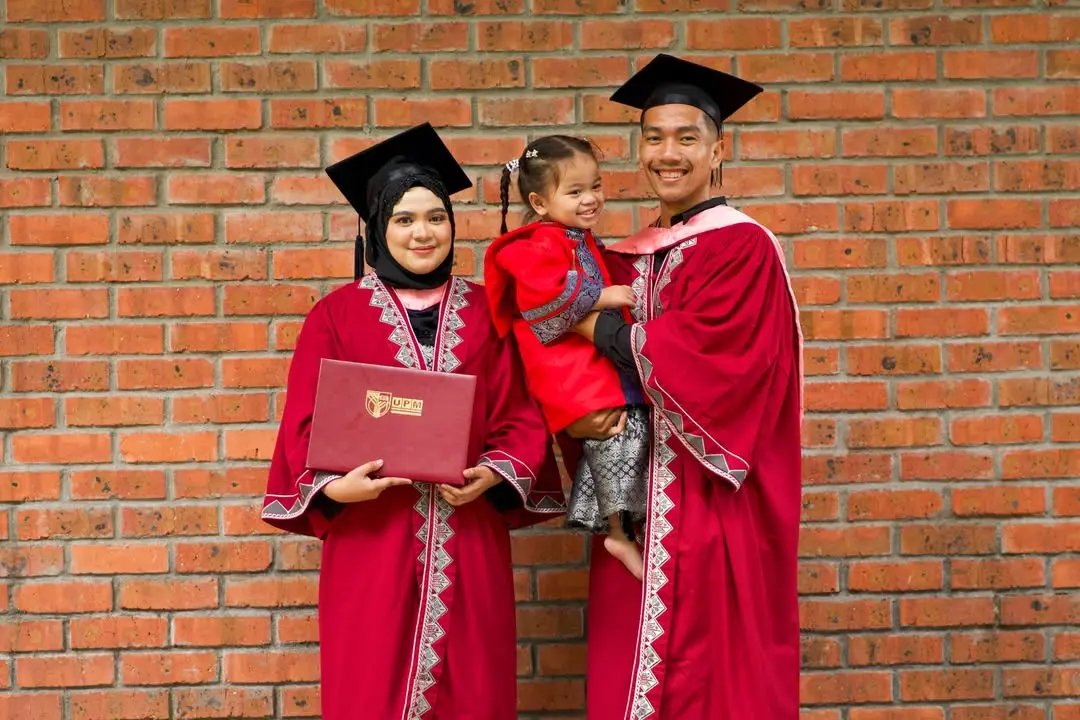 Pasangan graduan bergambar bersama anak kecil sambil memakai jubah konvokesyen universiti di hadapan dinding bata.