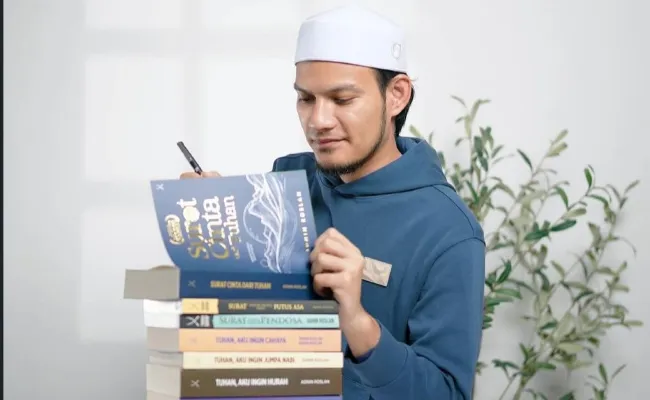 Seorang pemuda, kelihatan seperti Ustaz Adnin, sedang memegang dan melihat sebuah buku di atas timbunan buku.