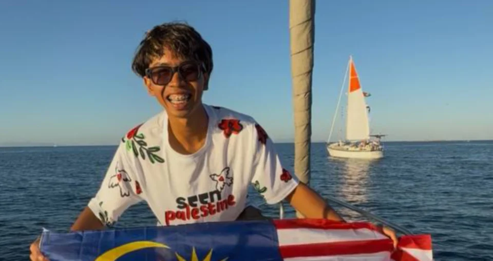 Muhammad Hareez Adzrami (Harogs), seorang pelajar UPSI, bergambar sambil memegang bendera Malaysia di atas kapal di tengah laut. Beliau memakai cermin mata hitam, braces, dan baju-T putih dengan cetakan bunga merah dan perkataan "SEEN Palestine" di bahagian hadapan, dengan sebuah kapal layar kelihatan di belakangnya.