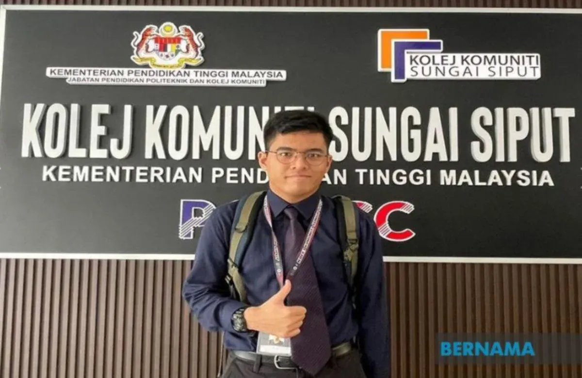 Seorang graduan Kolej Komuniti berdiri di hadapan papan tanda Kolej Komuniti Sungai Siput sambil menunjukkan isyarat ibu jari sebagai simbol kejayaan pendidikan
