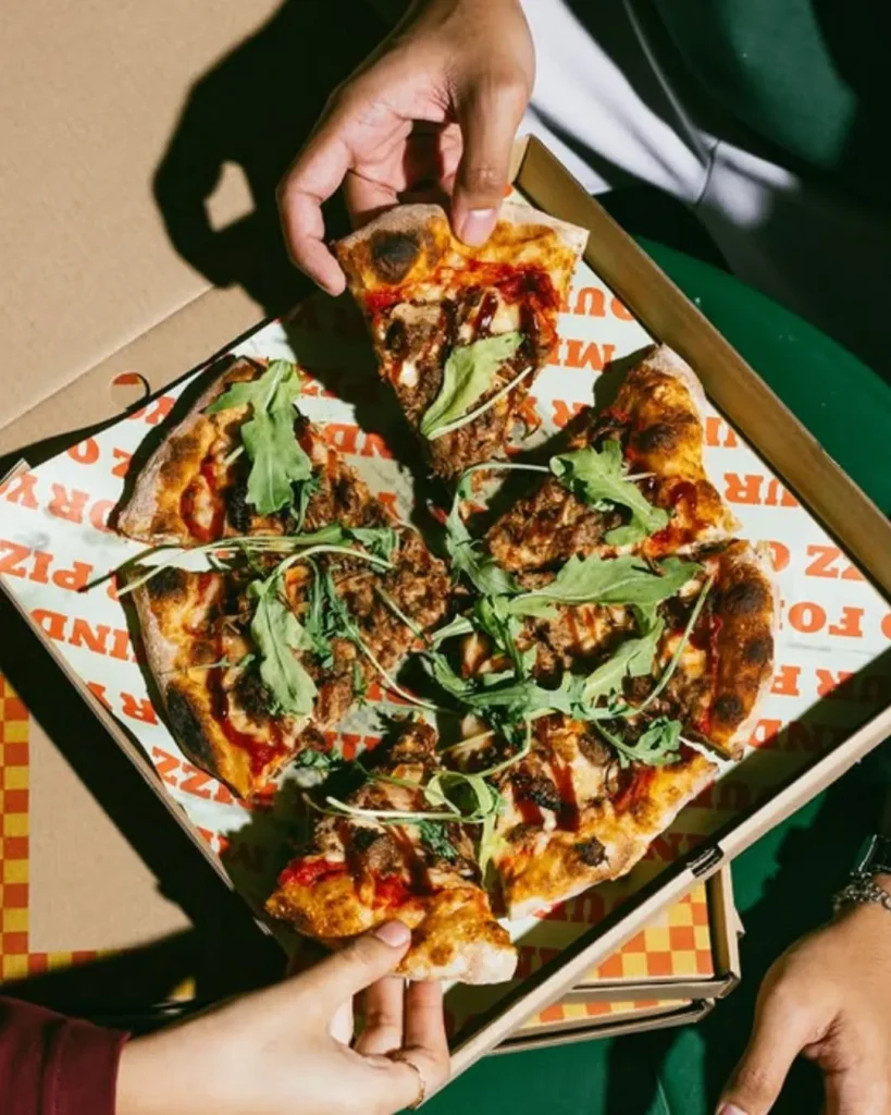 Beberapa tangan mengambil kepingan pizza Mokky’s Pizza dengan topping daging dan daun arugula di dalam kotak pizza