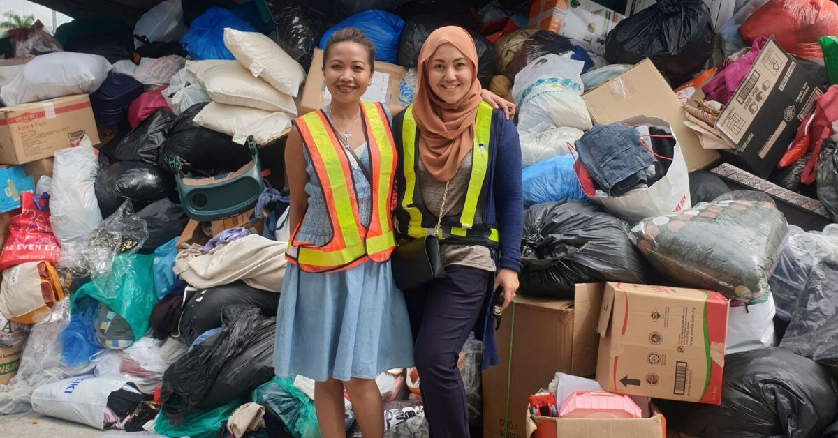 Dua wanita memakai vest keselamatan berdiri di hadapan longgokan sisa fabrik dan pakaian untuk proses kitar semula tekstil di Malaysia