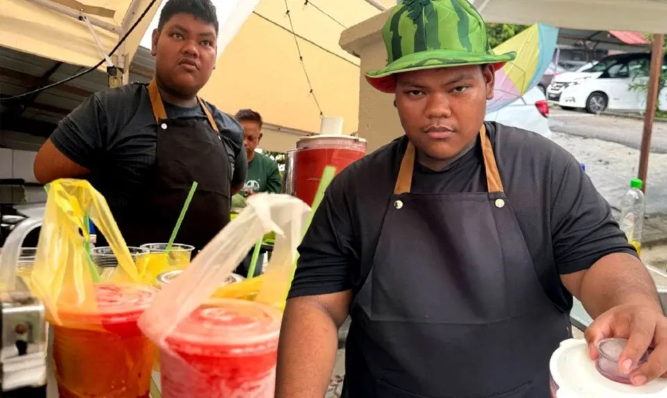 Seorang penjual minuman jalanan memakai apron hitam, muncul dua kali dalam gambar, salah satunya memakai topi bertema tembikai, sedang menghidangkan minuman berwarna-warni dengan cawan merah dan kuning di hadapan.
