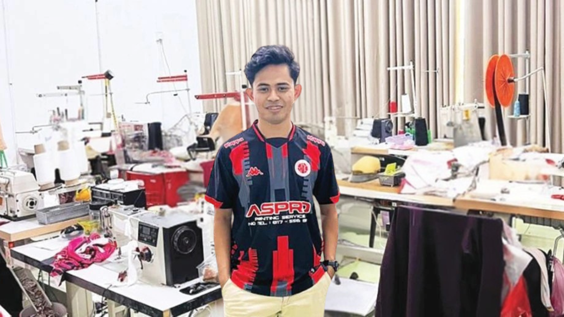Akmal usahawan muda berdiri dalam kilang cetak baju dengan mesin jahit dan pekerja di belakang