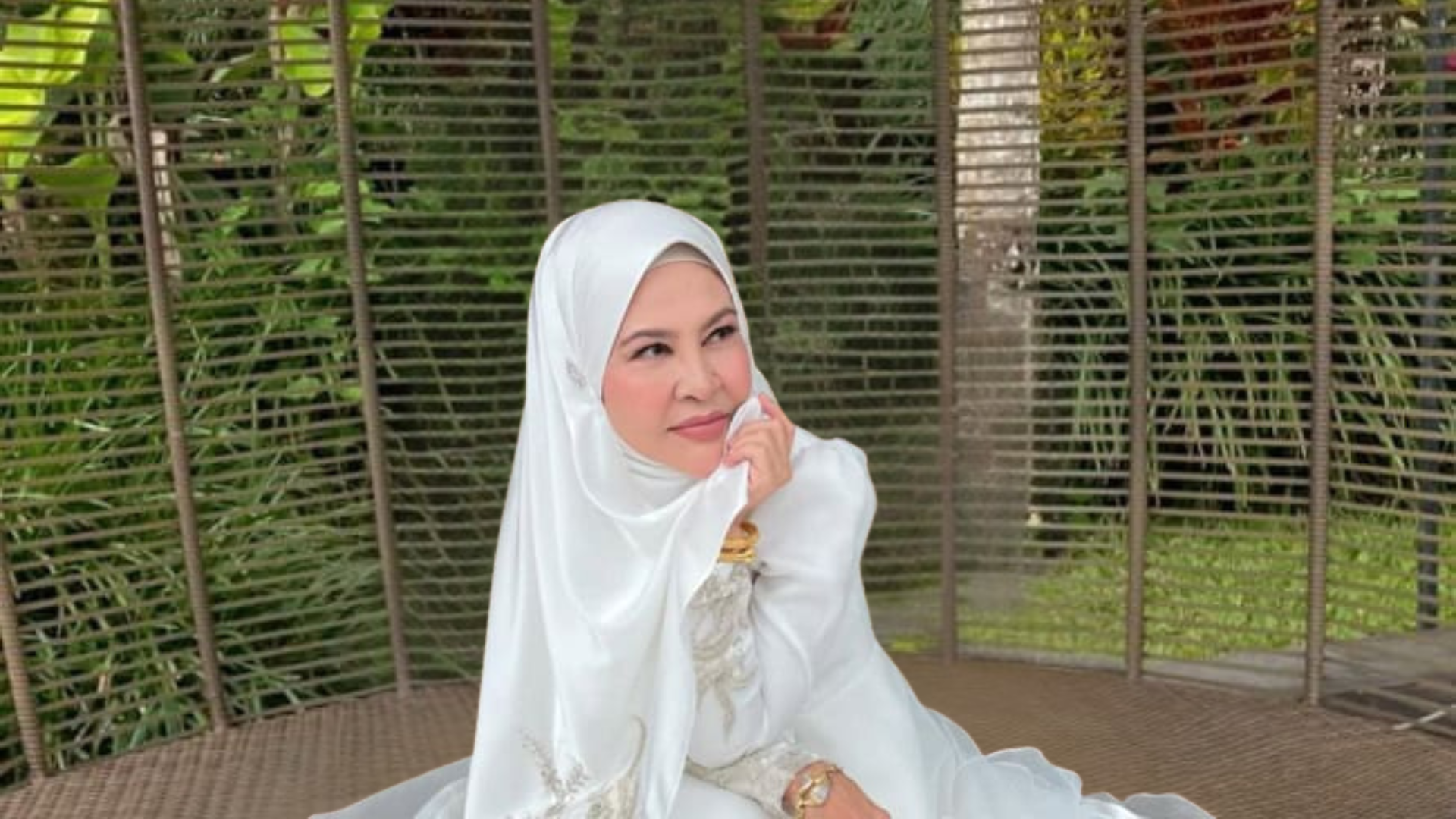 Wanita berhijab putih bergaya santai di kerusi rotan berlatar suasana taman hijau